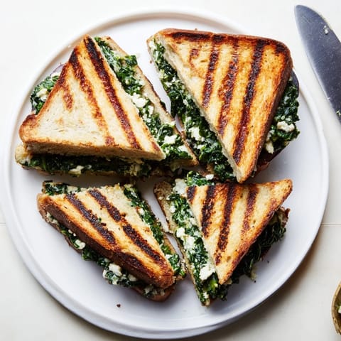 A vibrant Feta & Spinach Stuffed Grilled Cheese rests on a white plate, showcasing crispy sourdough and flecks of green spinach.